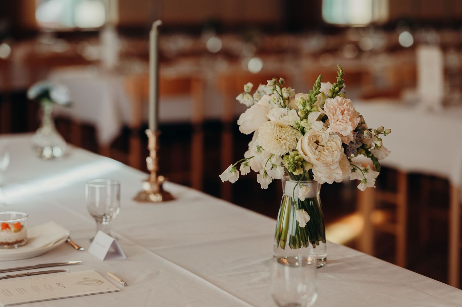 Delicate white and blush floral centerpiece in glass vase on white linen wedding reception table.