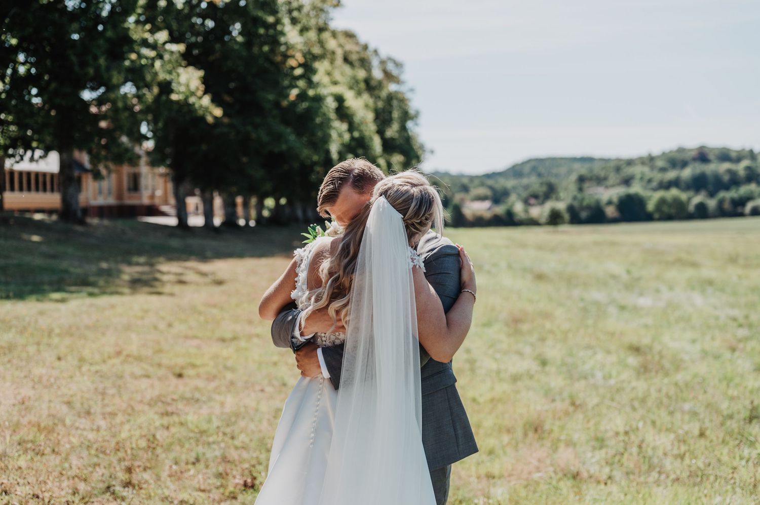 Bride and groom embracing in a sunny green field on their wedding day, with trees and hills in the background.