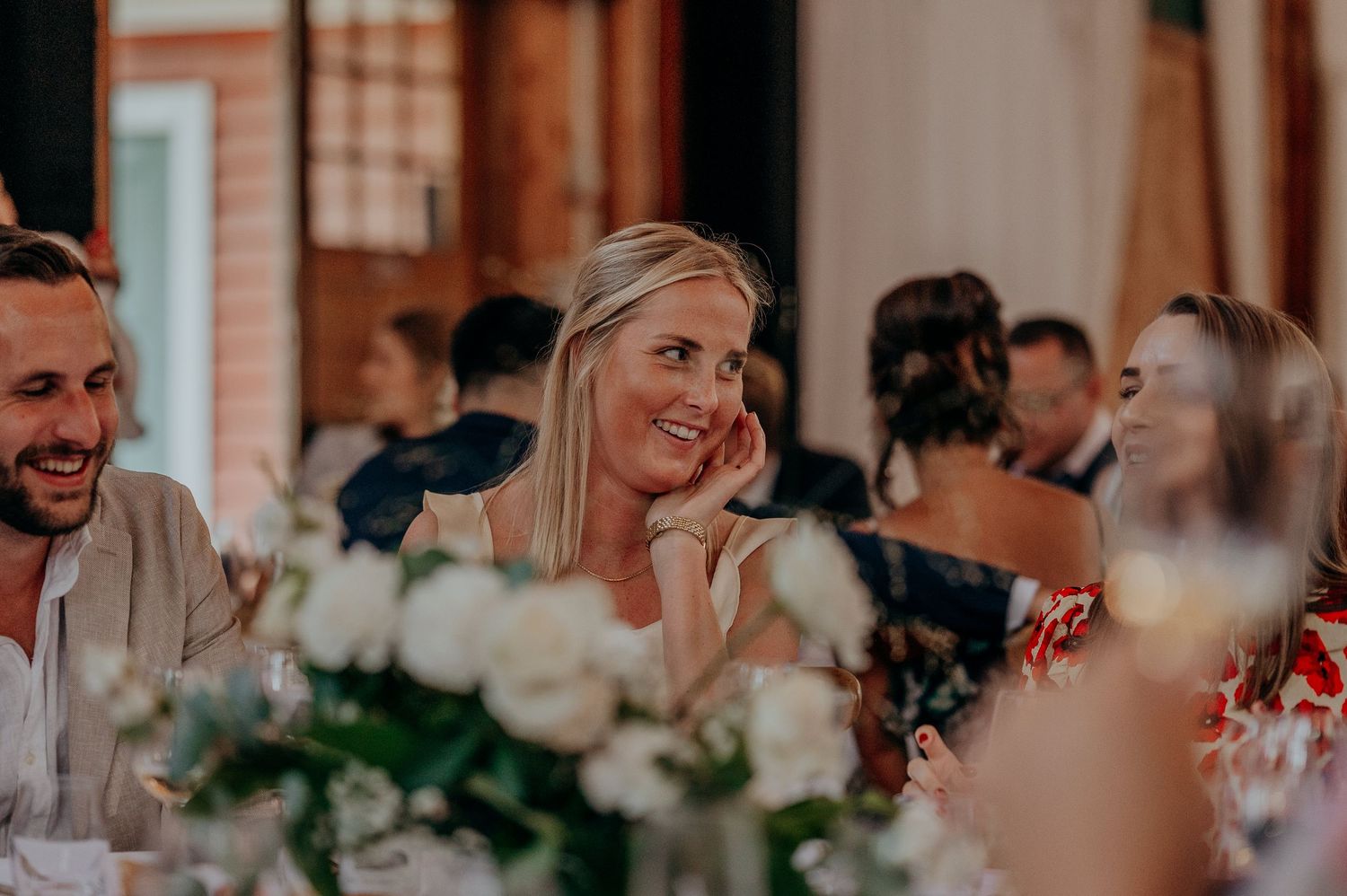 Smiling blonde woman at elegant wedding reception dinner, surrounded by white floral centerpieces and guests.