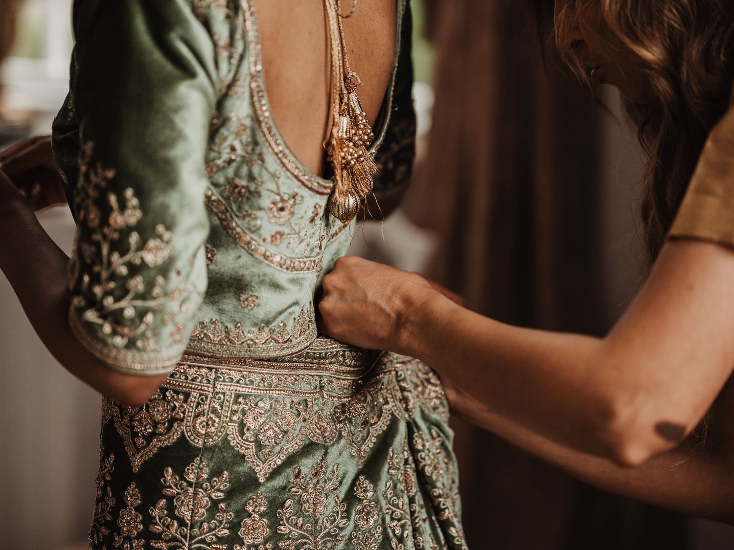 Close-up of a bride in a sage green embroidered lehenga being fastened by her mom.