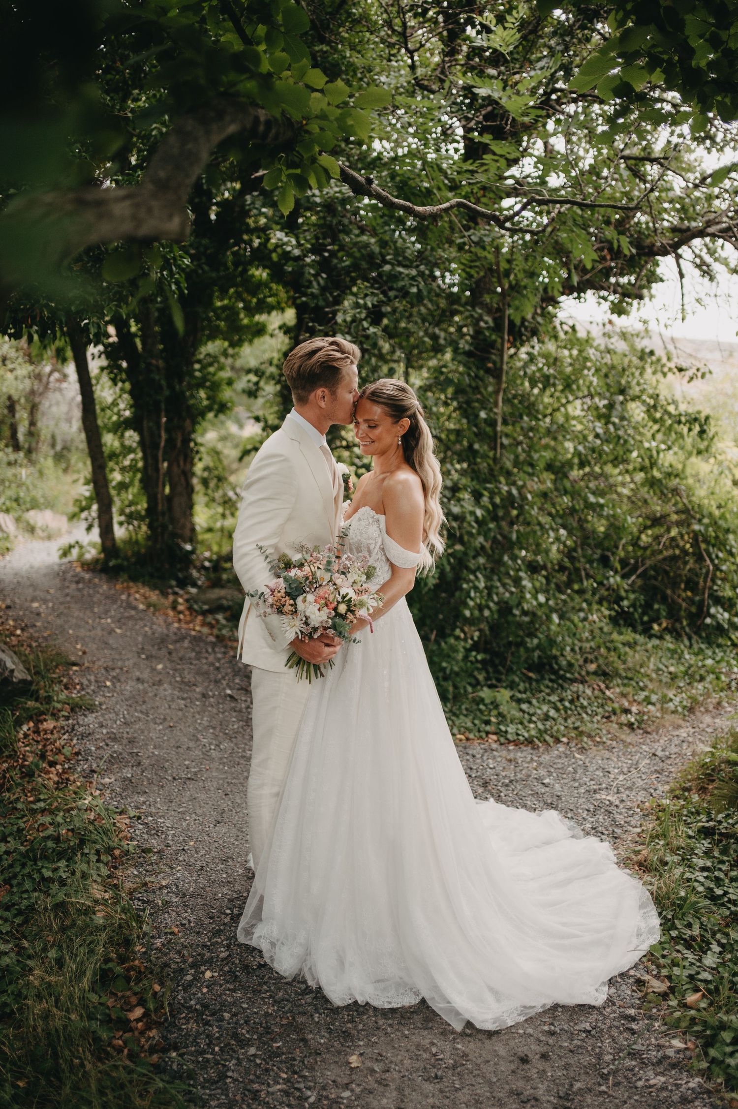 Bride and groom share a romantic moment on a garden path, bride holding a pink bouquet in a flowing off-shoulder gown.