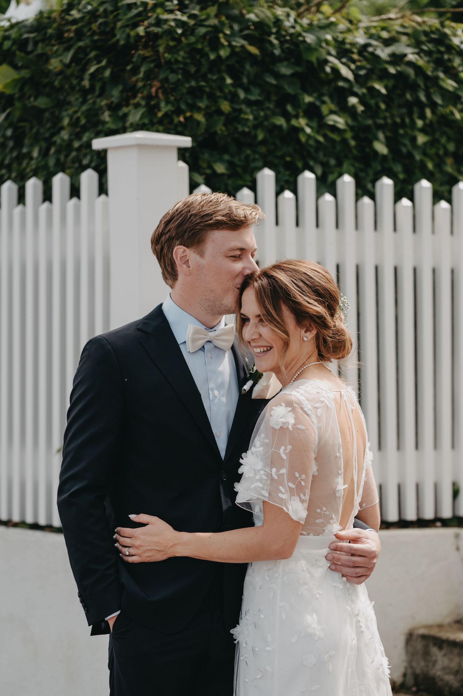 Groom in black suit kisses bride in lace wedding dress by white picket fence outdoors.