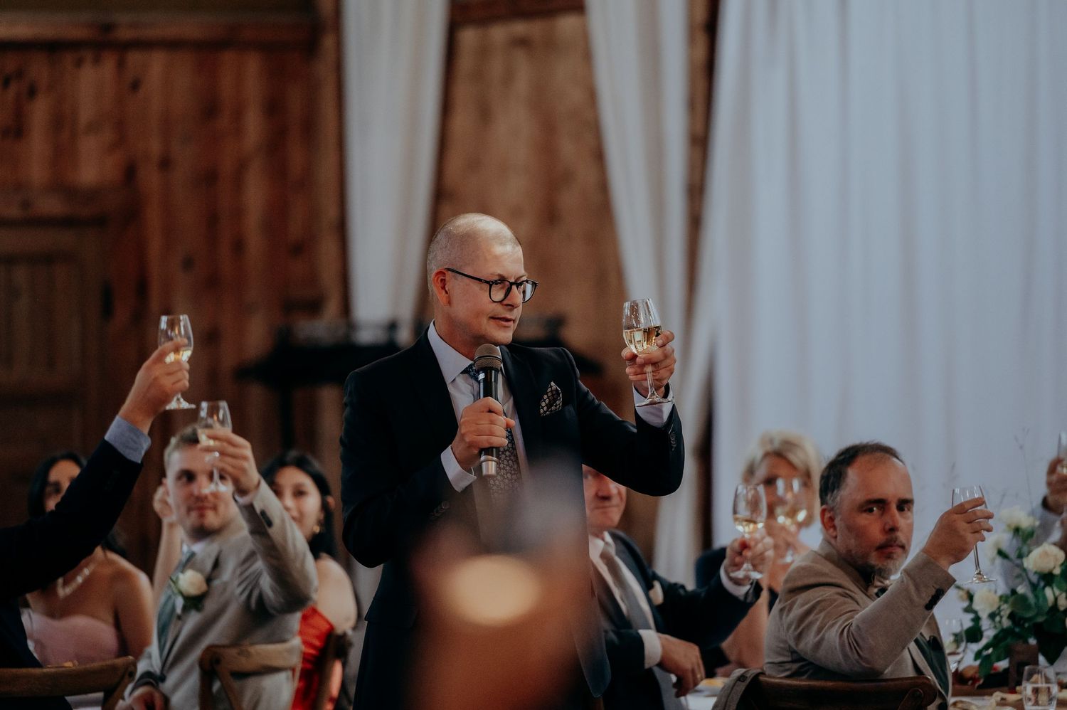 Man giving a toast at a wedding reception, raising a champagne glass while guests celebrate in a rustic barn venue.