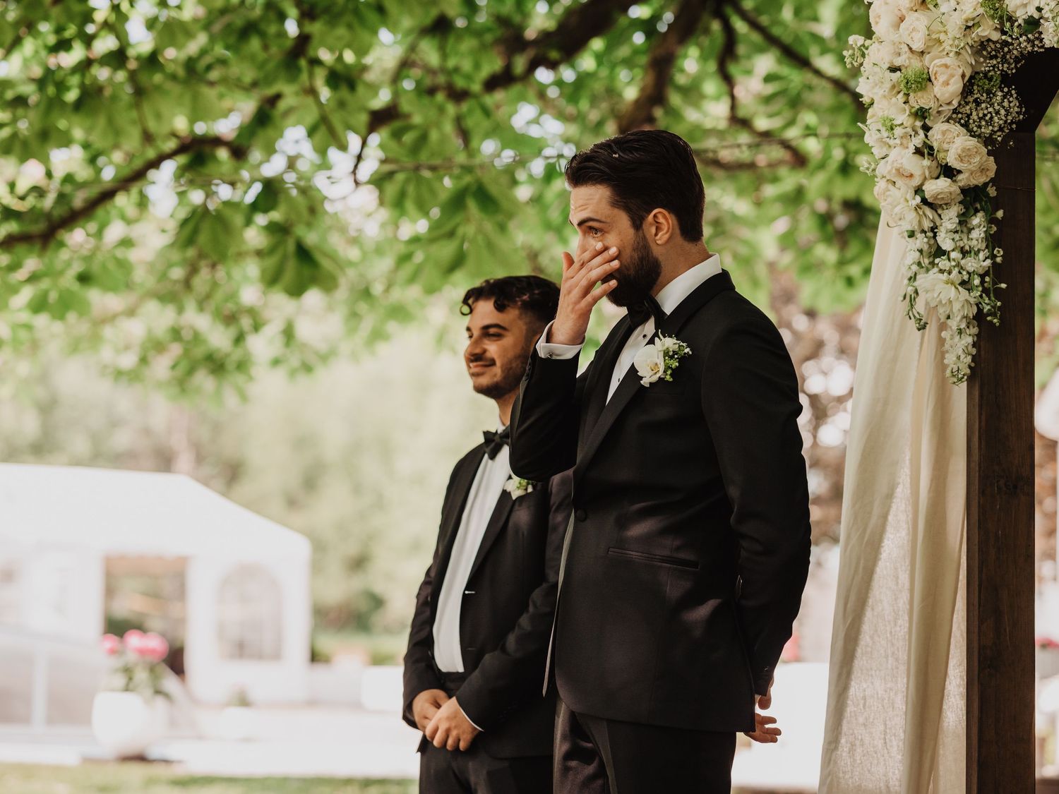 Two grooms in black tuxedos share an emotional moment at outdoor wedding ceremony surrounded by lush greenery and floral arch.