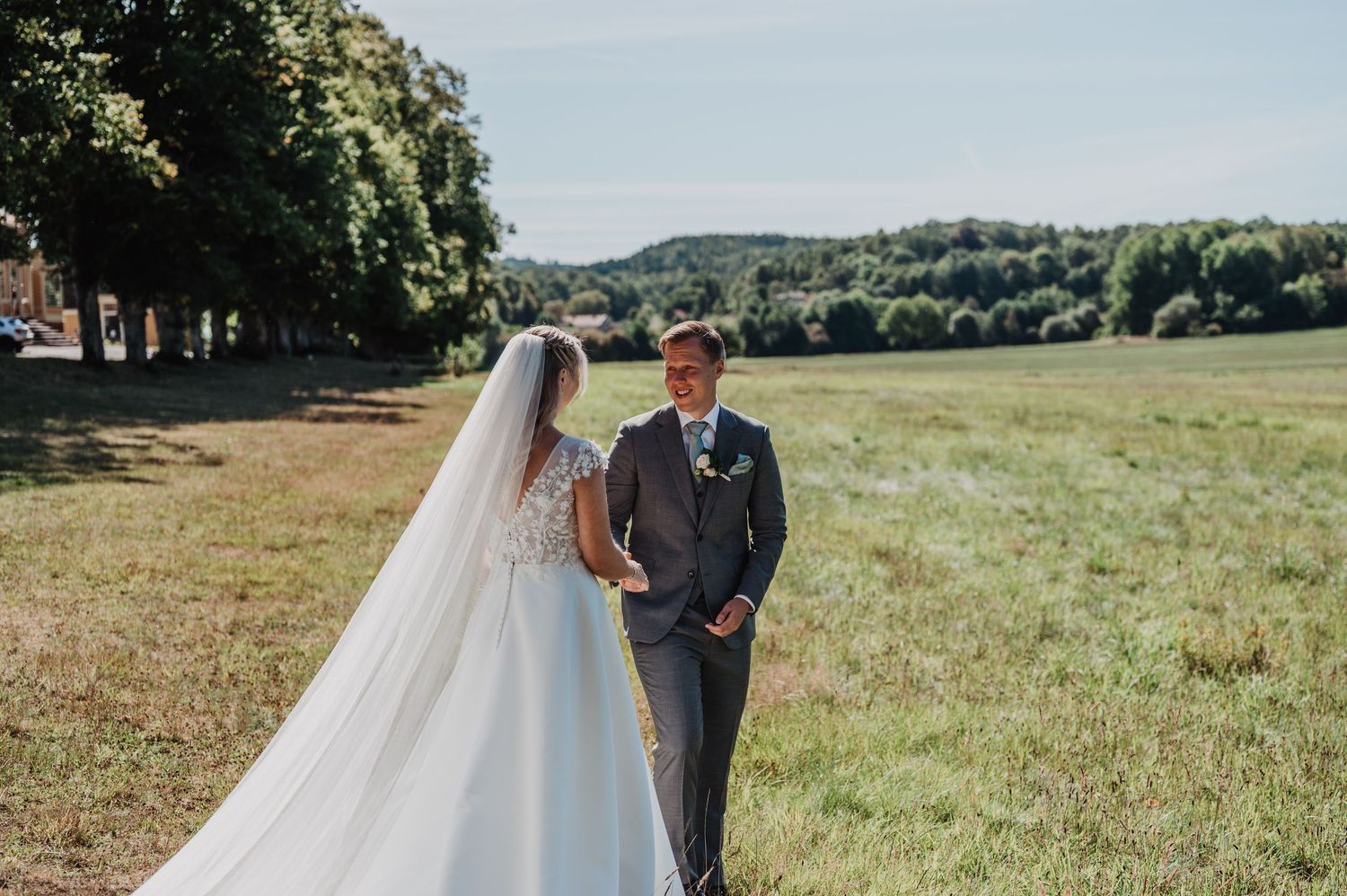 Bride in flowing white gown and veil faces groom in gray suit during outdoor first look on sunny green field.