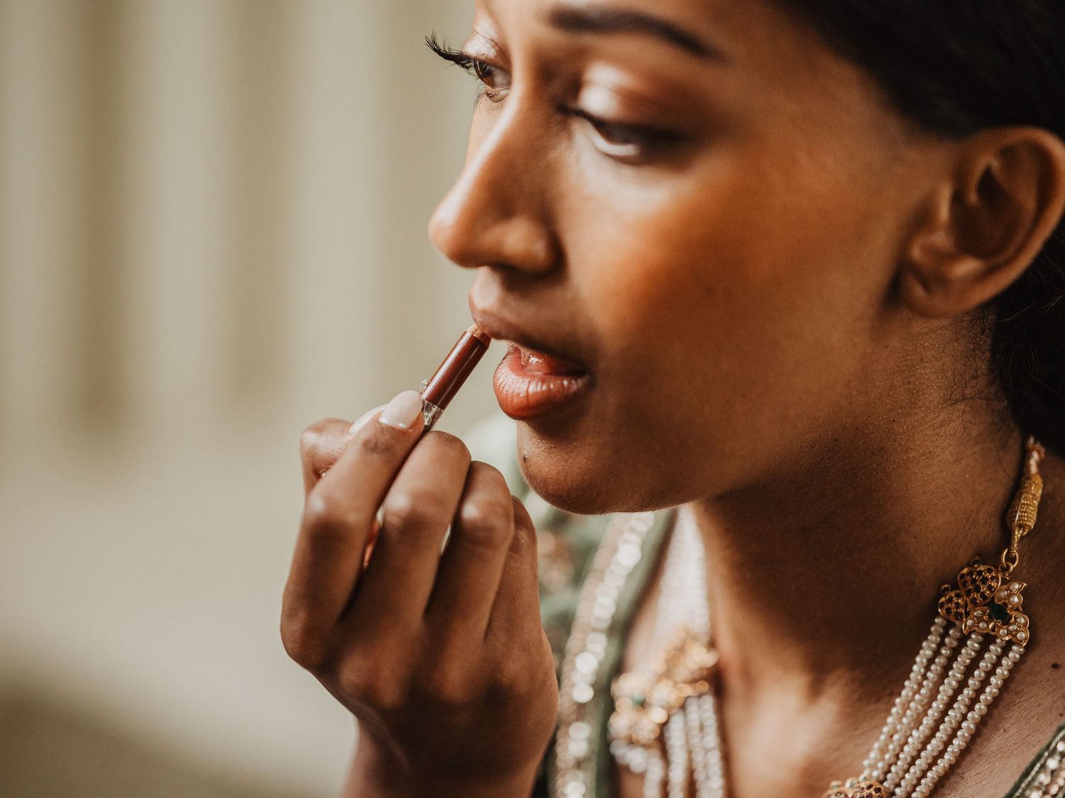 Close-up of a South Asian bride carefully applying lipstick while wearing elegant gold jewelry.