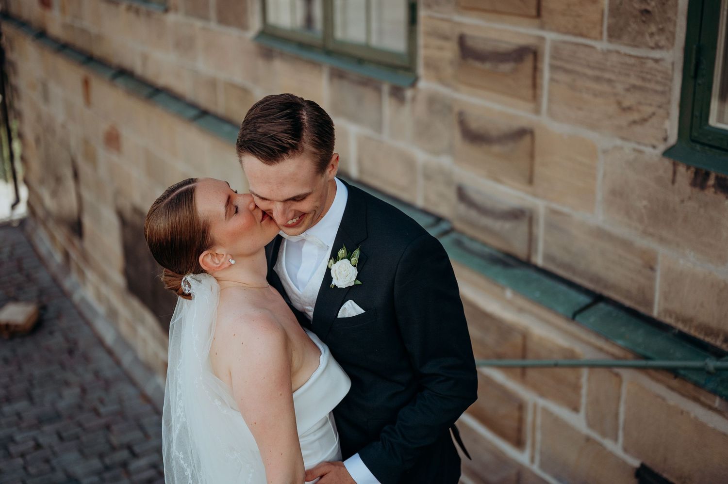 A bride and groom share a romantic kiss against a rustic sandstone wall on their wedding day.