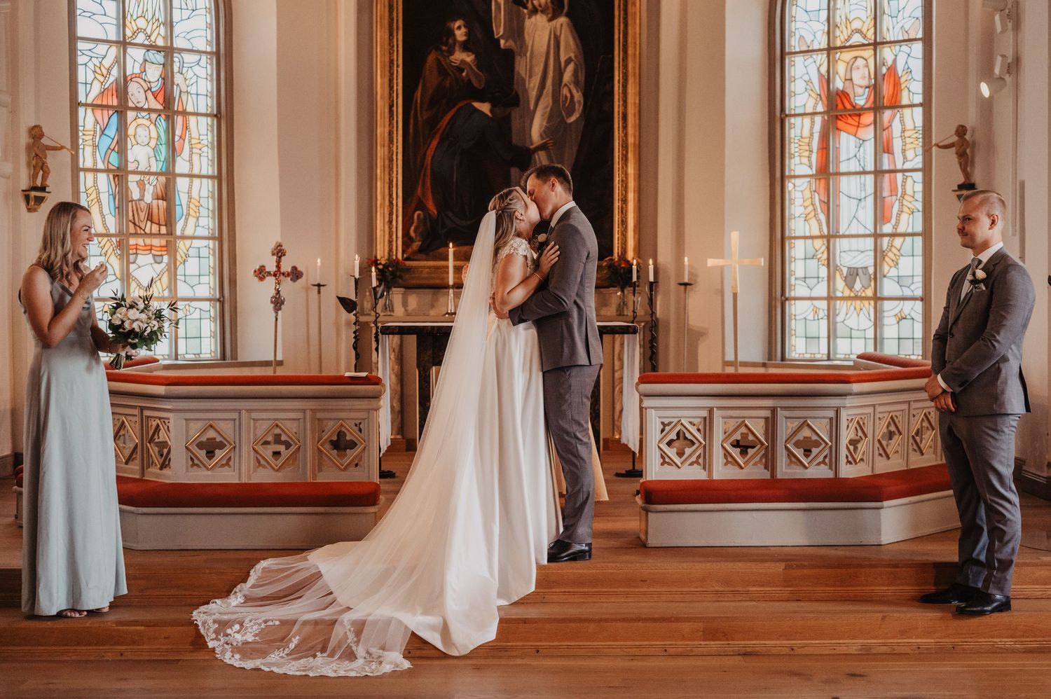 Bride and groom share first kiss at church altar flanked by bridesmaids and groomsmen with stained glass windows.