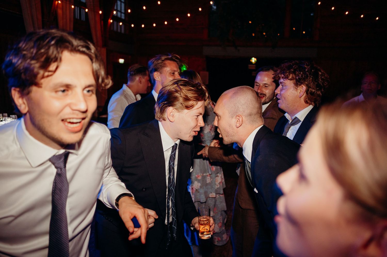 Male wedding guests share a lively conversation under string lights at an evening wedding reception.