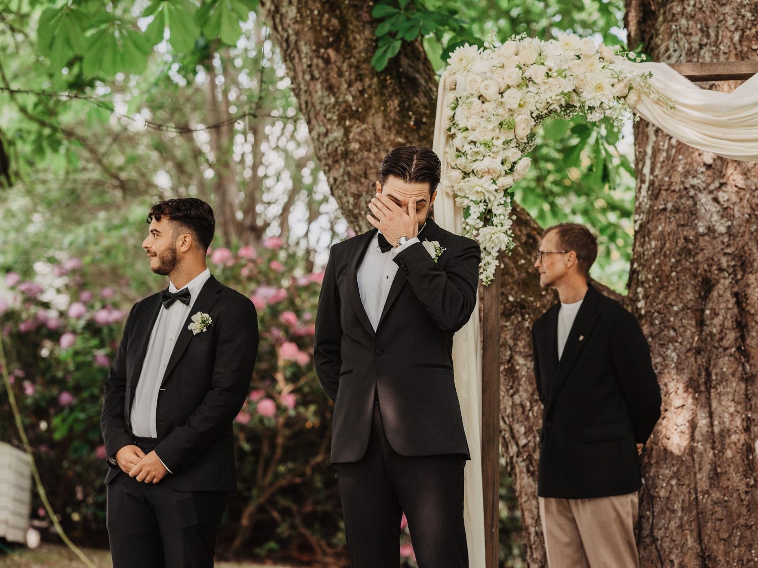 Emotional groom wiping tears at outdoor wedding ceremony with groomsman and officiant beside floral arch.