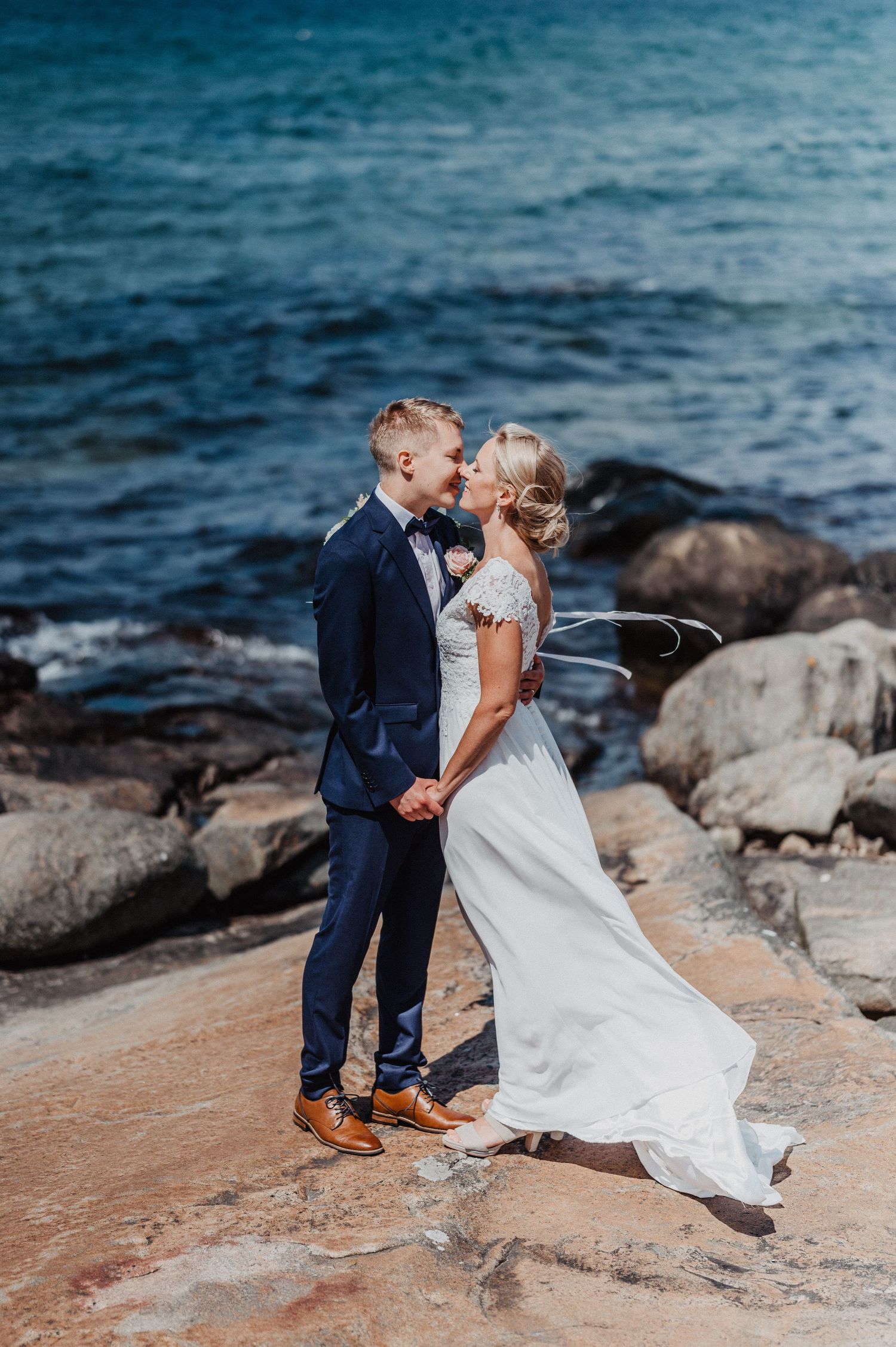 Bride and groom kissing on rocky shoreline with blue ocean waves in the background.
