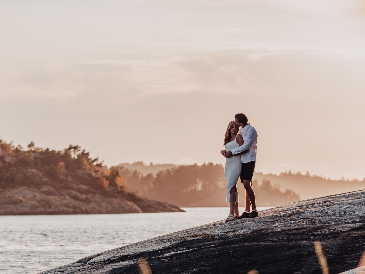 Couple embracing romantically on rocky shoreline at golden sunset with serene water and forest backdrop.