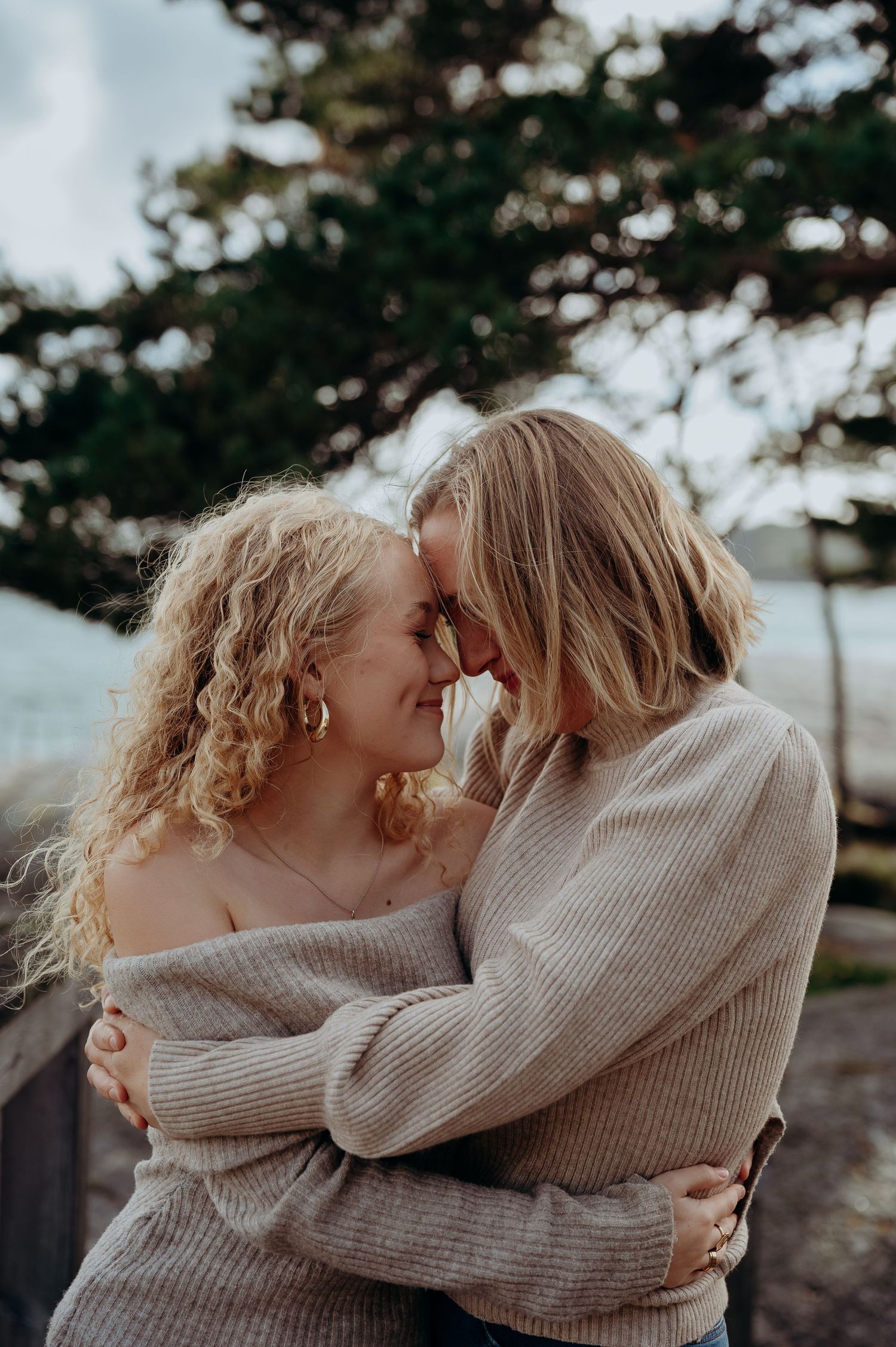 Two women hugging closely and touching foreheads in an affectionate outdoor coastal setting.