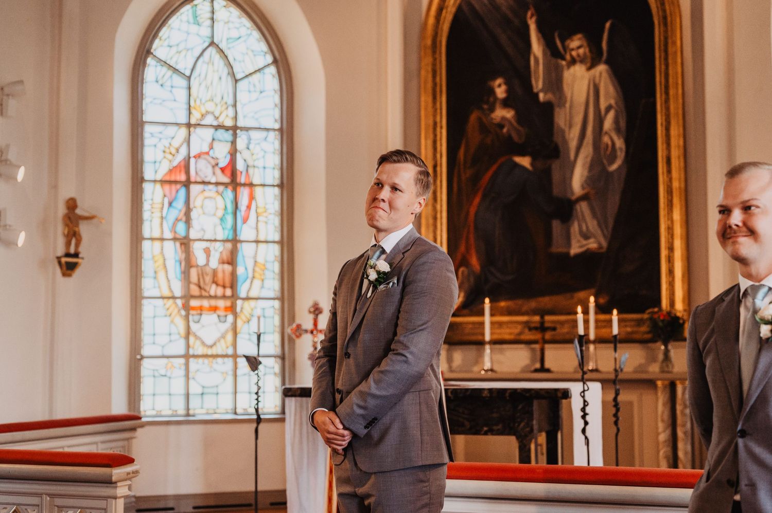 Groom in gray suit stands at church altar awaiting bride, with stained glass windows and religious painting behind him.