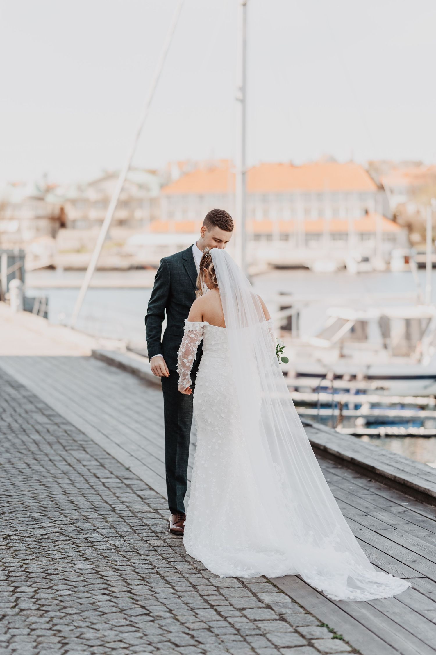 Bride and groom share a romantic moment on a cobblestone harbor dock, with sailboats and buildings in the background.
