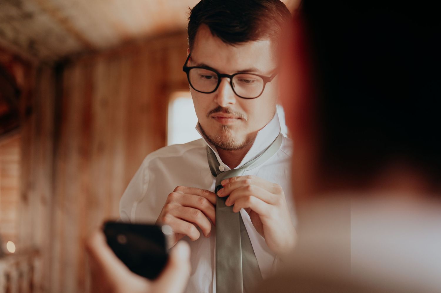 Groom adjusting his tie while checking his reflection on a phone screen before the wedding ceremony.