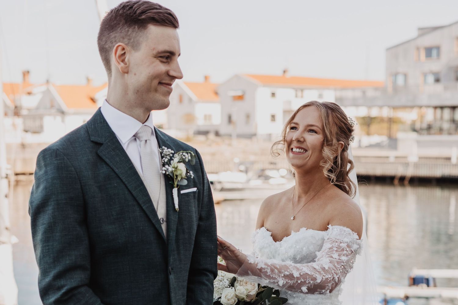 A smiling bride in a lace gown and groom in a grey suit pose together at a waterfront harbor on their wedding day.