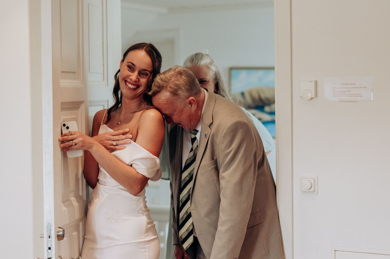Bridesmaid in pink dress smiles while showing the bride to older dad in suit at doorway.