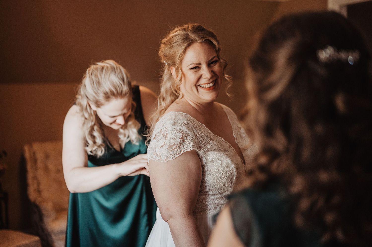 Bridesmaid helping bride with wedding dress while bride smiles radiantly in lace gown.