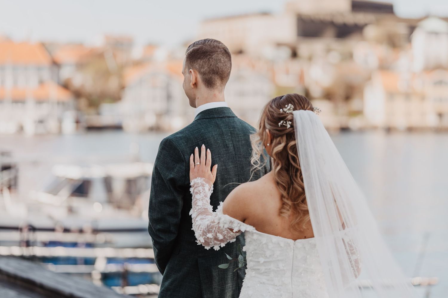 Bride and groom stand together overlooking a marina, bride wearing lace gown and veil on their wedding day.