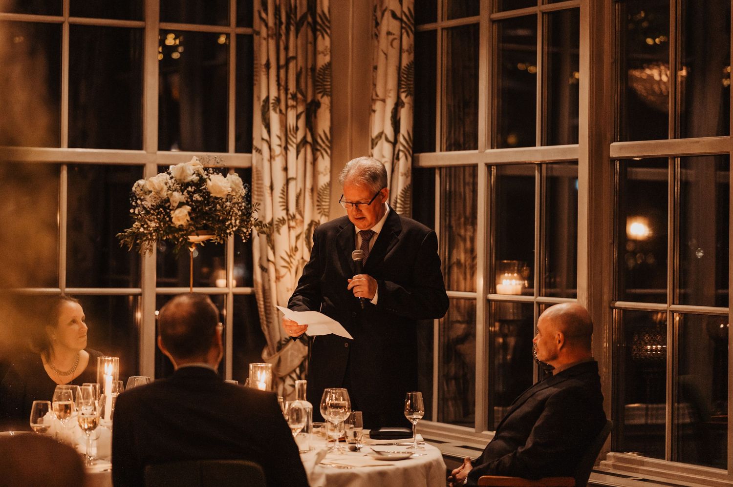 Man in suit giving a speech at an elegant candlelit wedding reception dinner with floral centerpieces.