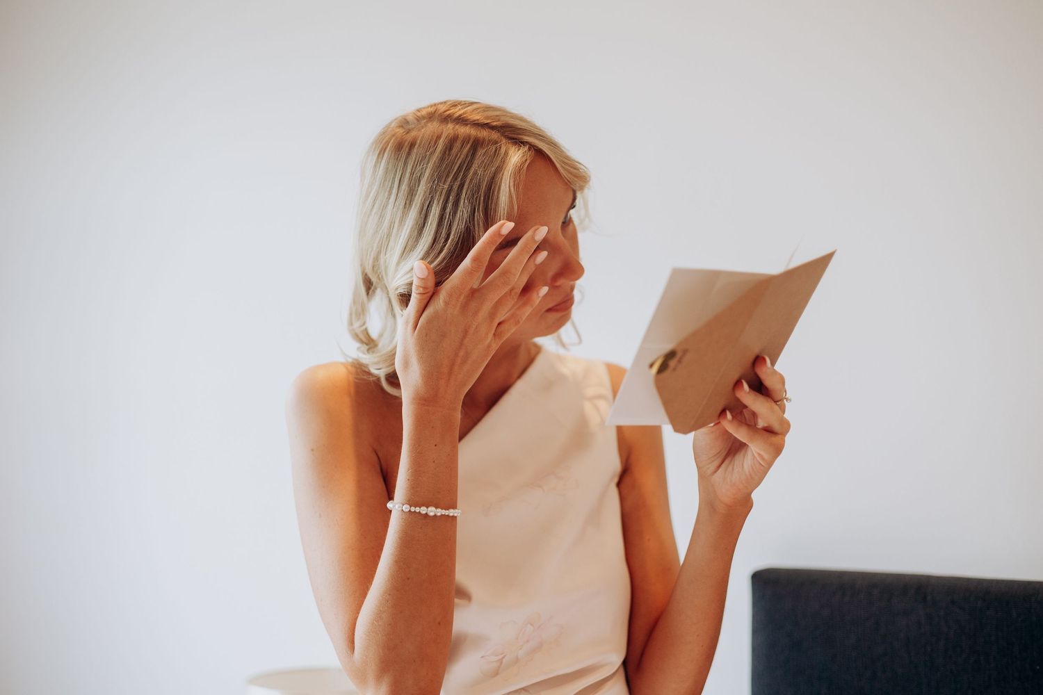 Bridemaid in sleeveless top reading a card while touching her face, seated against a white background.