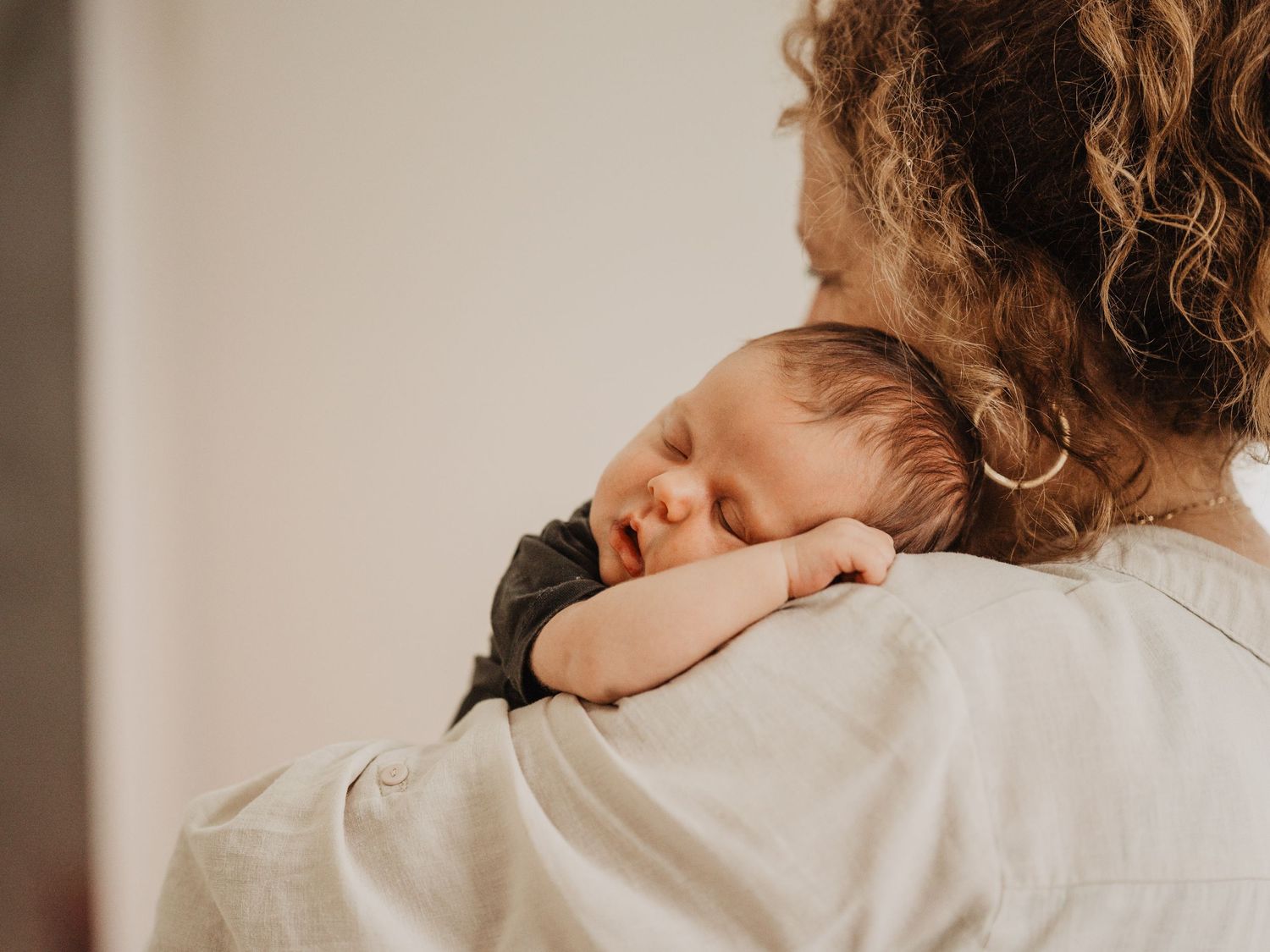 A curly-haired mother gently holds her sleeping newborn baby on her shoulder in a soft, natural light setting.