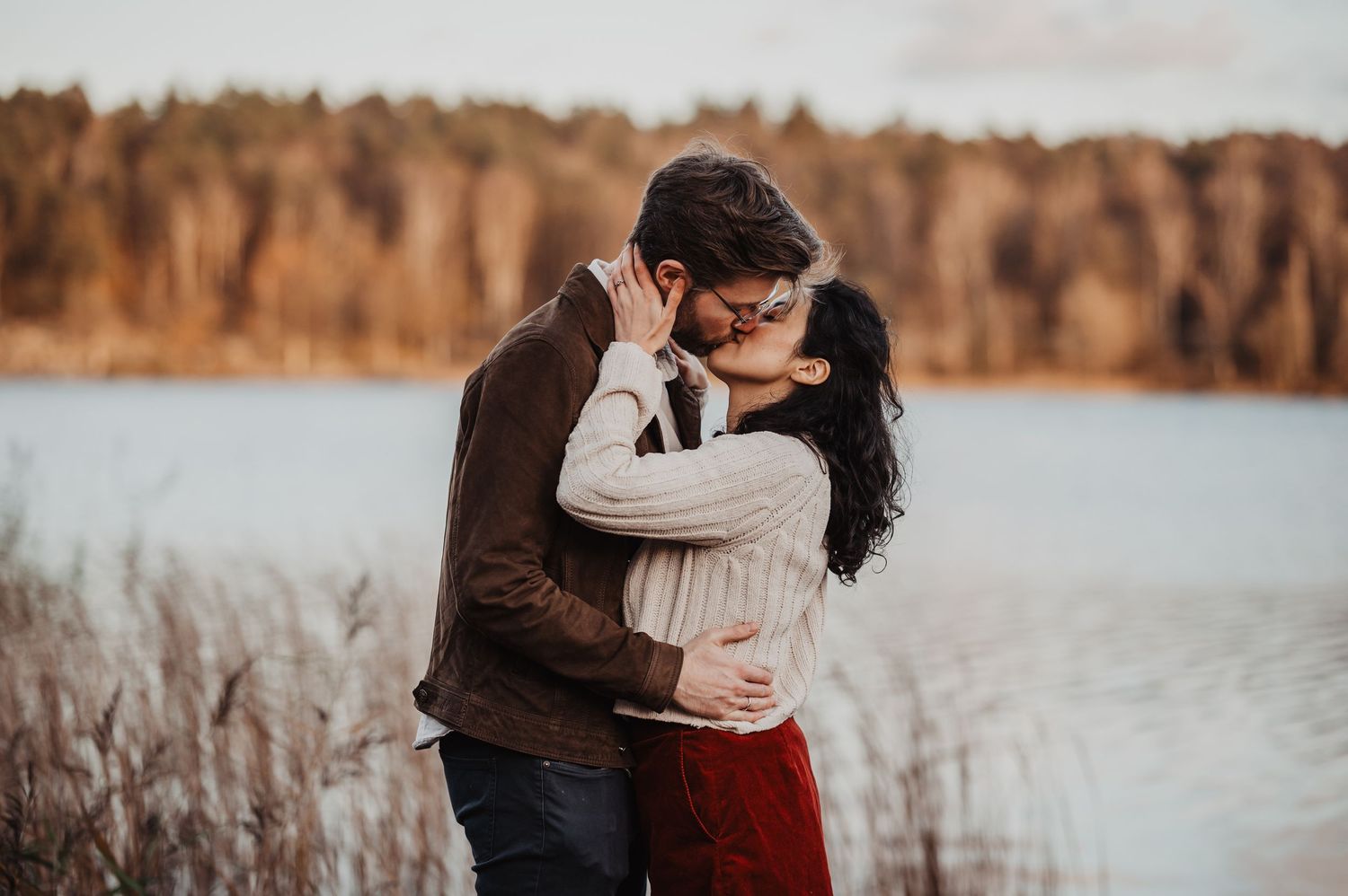 Couple sharing a tender kiss near a tranquil lake surrounded by autumn foliage and tall grasses.