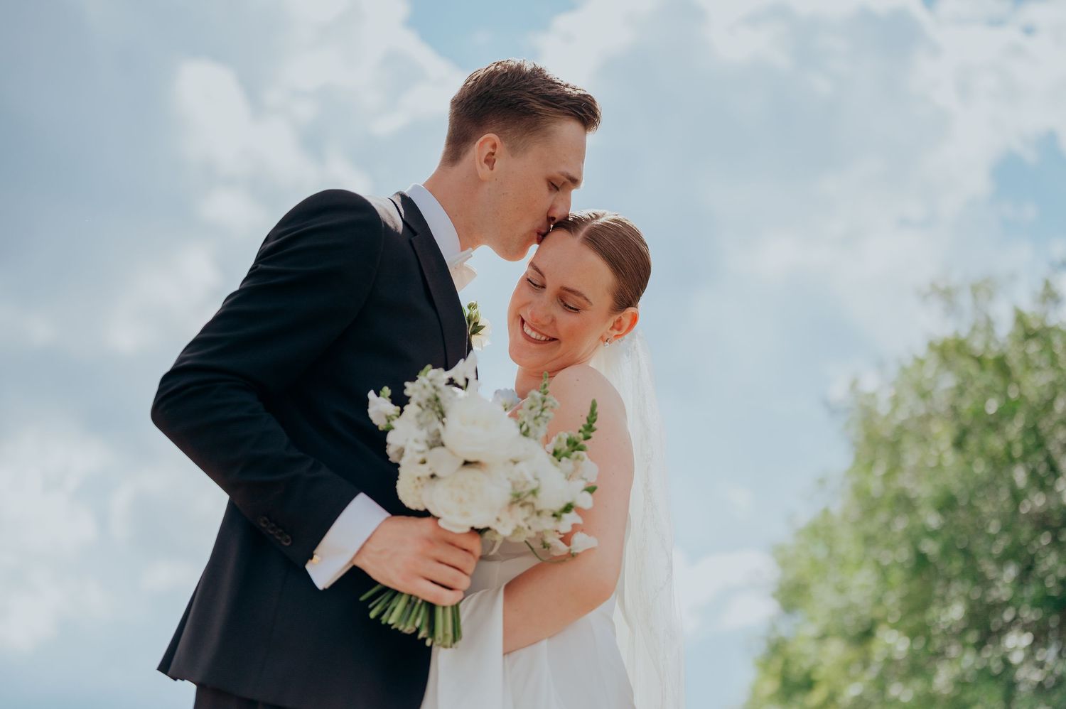 Joyful bride and groom embracing outdoors, holding white bouquet against a bright blue sky.