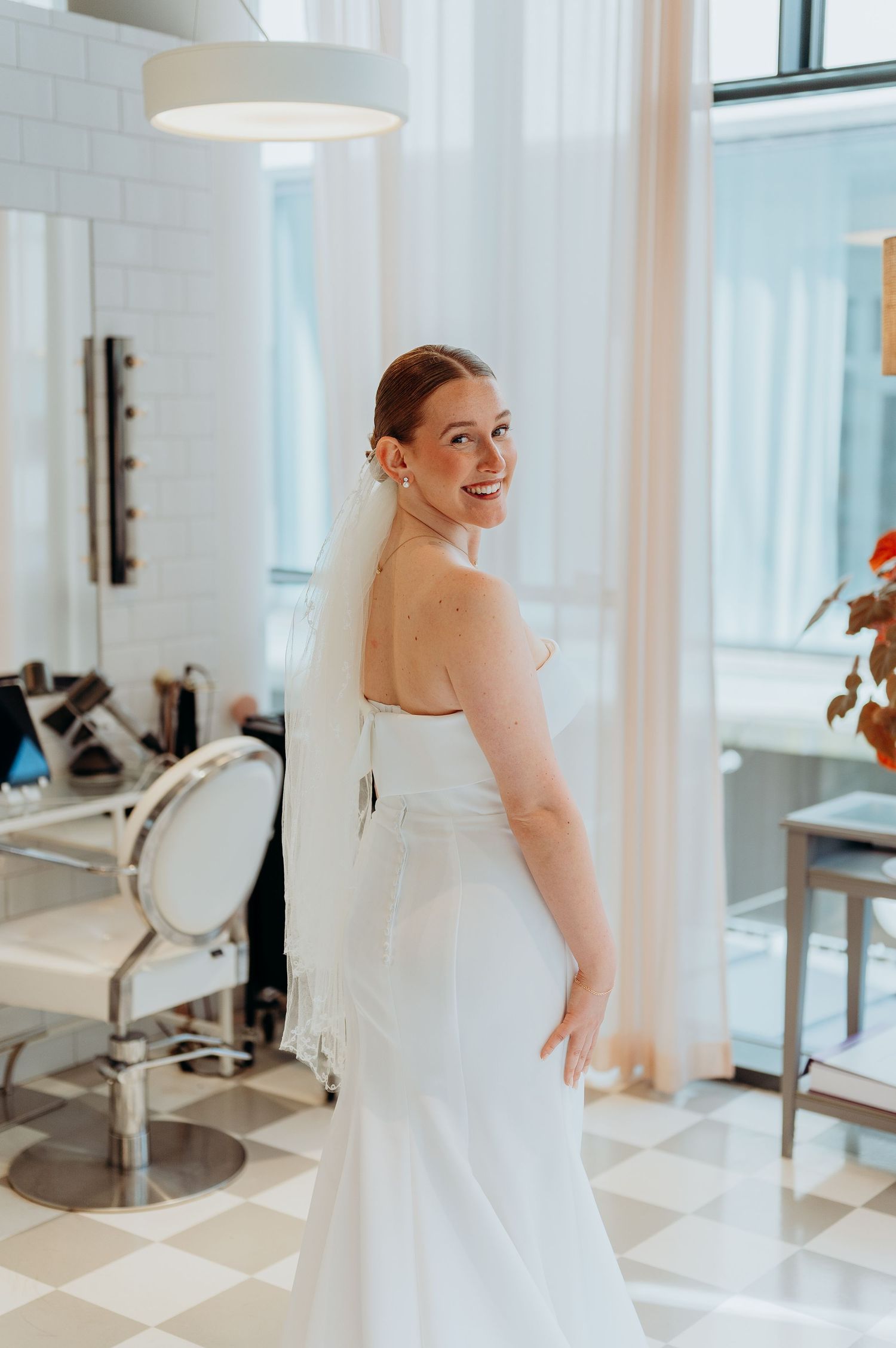 Radiant bride in strapless white wedding gown smiles over shoulder in bright bridal salon with checkered floor.
