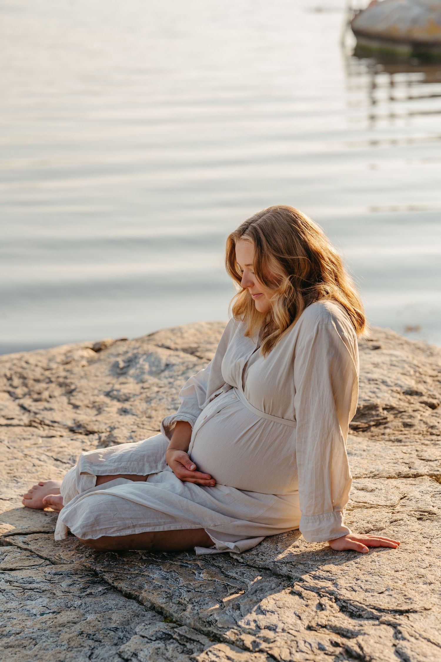 Pregnant woman in white dress sitting peacefully on rocks beside serene water at sunset.