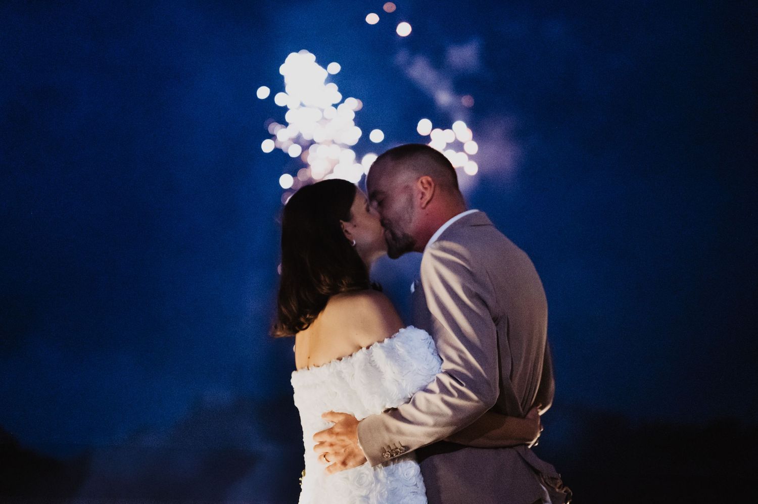 Newlyweds sharing a romantic kiss beneath a spectacular fireworks display against a dark blue sky.