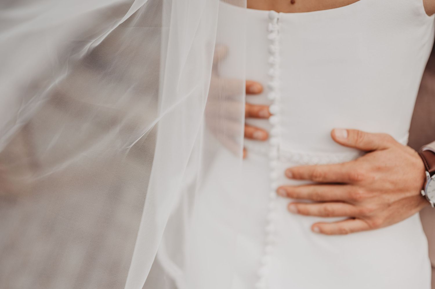 Close-up of grooms hand on brides button-back wedding dress with flowing veil.