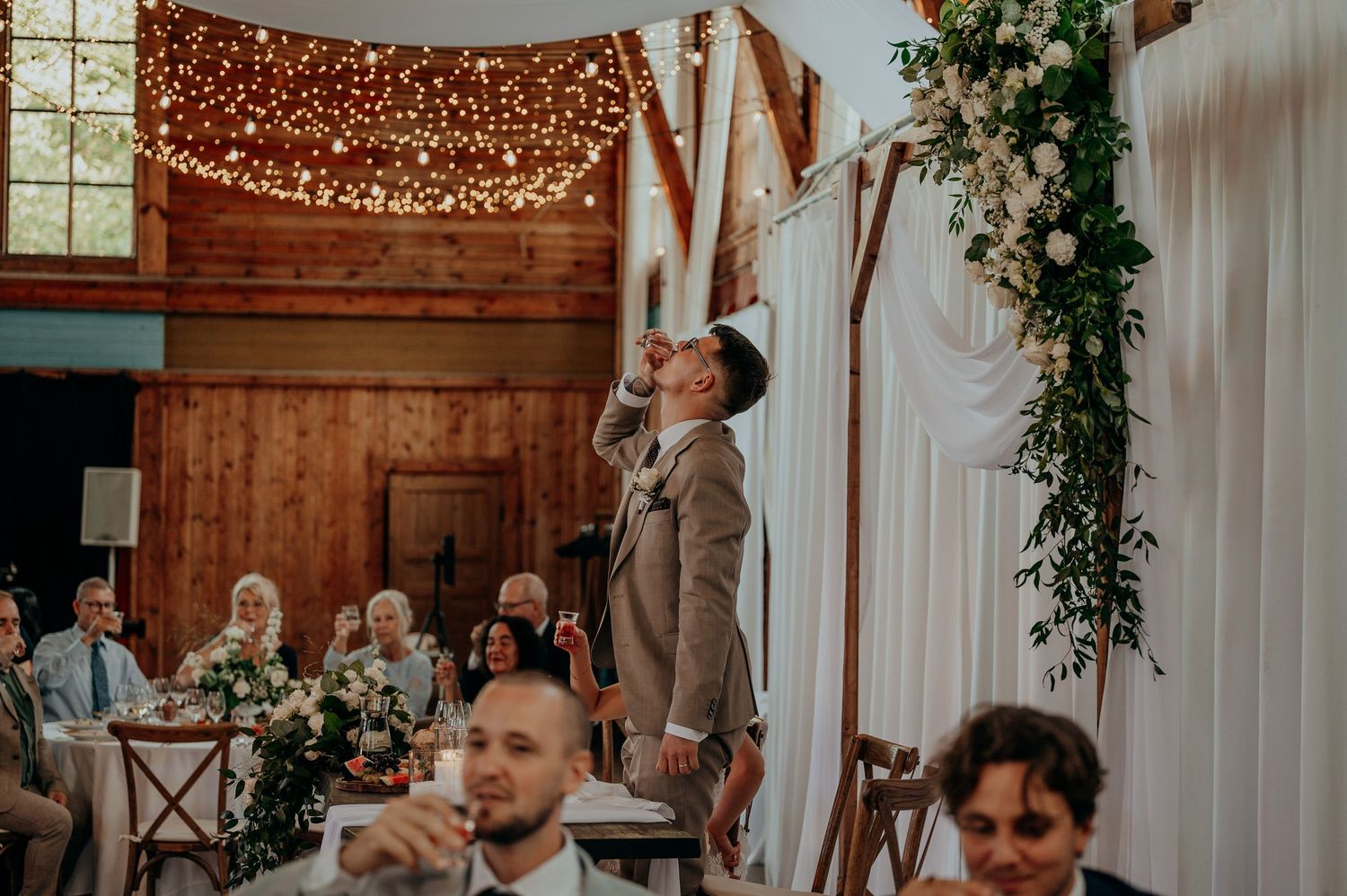 Groom taking a celebratory shot in rustic barn wedding venue with fairy lights.