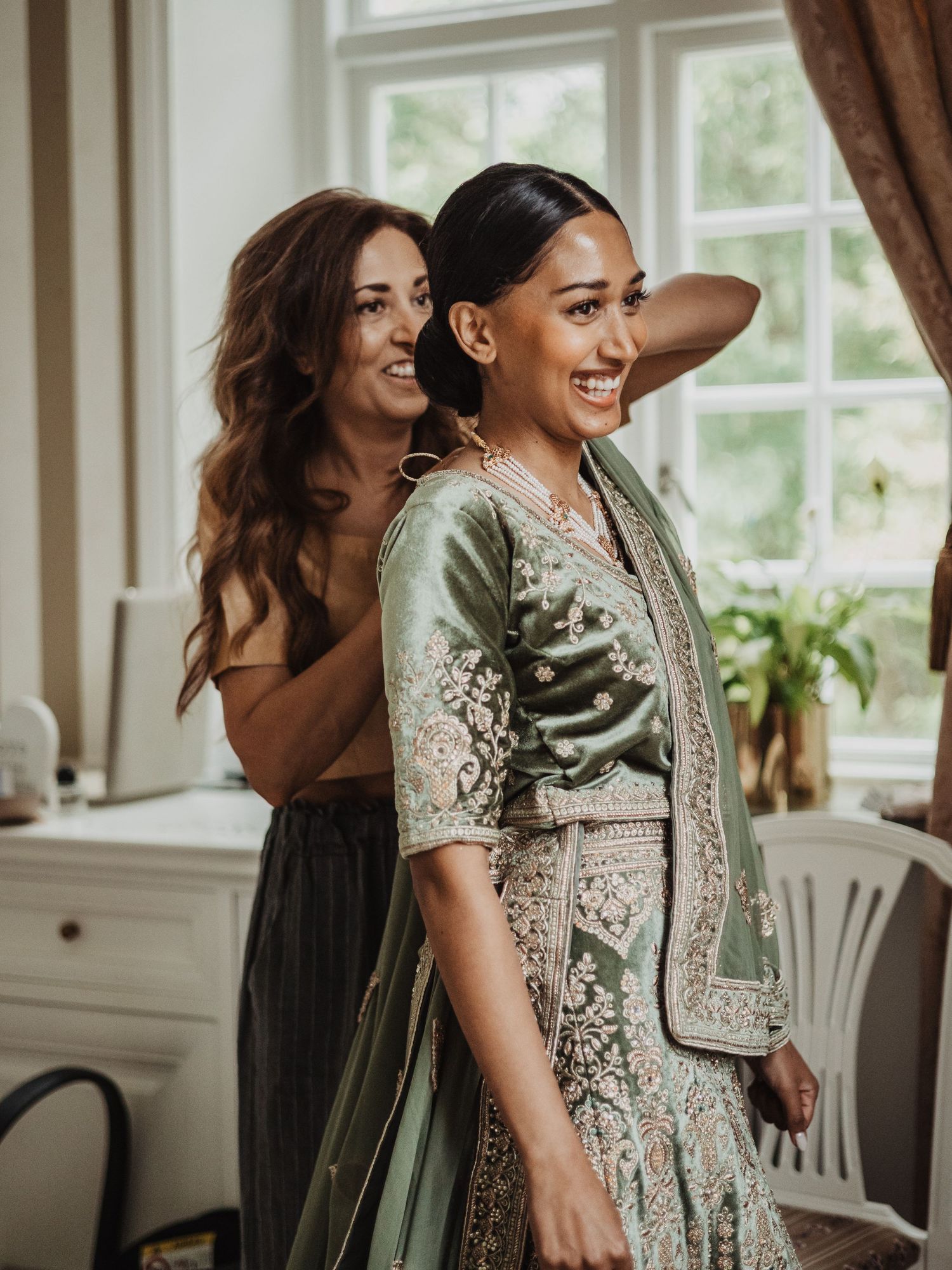 A smiling bride in a green embroidered lehenga laughs with her mom during wedding preparations.