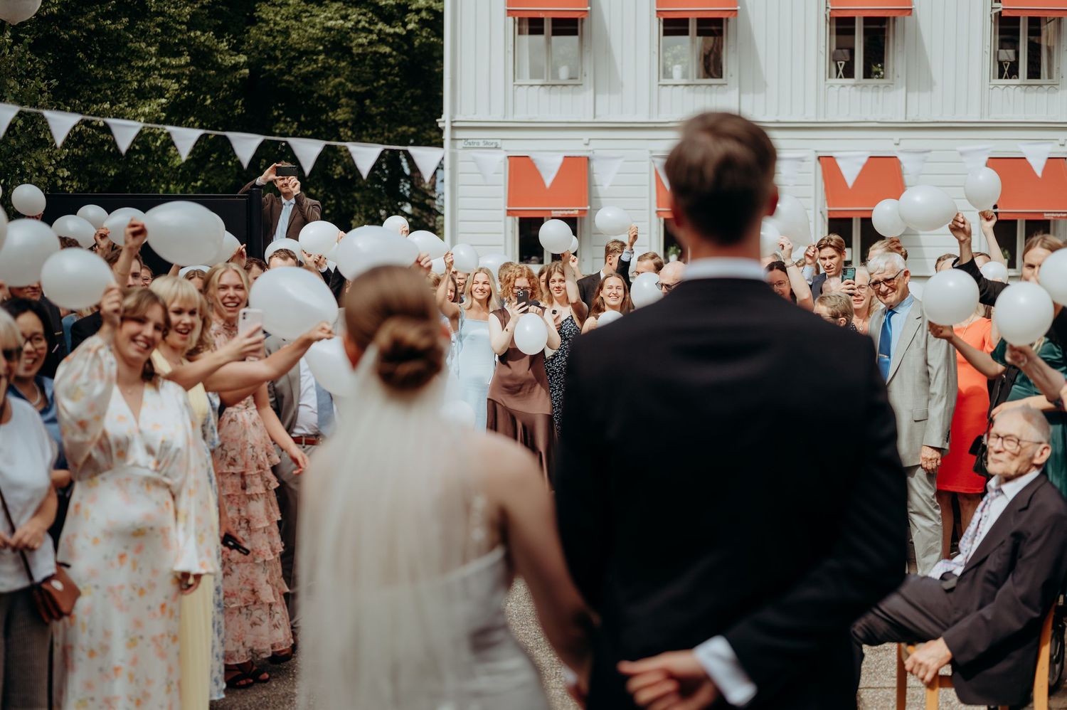 Wedding couple faces guests holding white balloons outdoors at a festive celebration near a decorated building.
