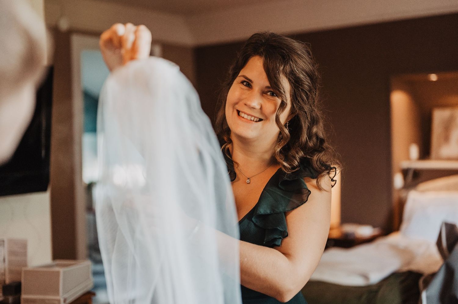 Smiling bridesmaid in green dress holds up white wedding veil in hotel room.