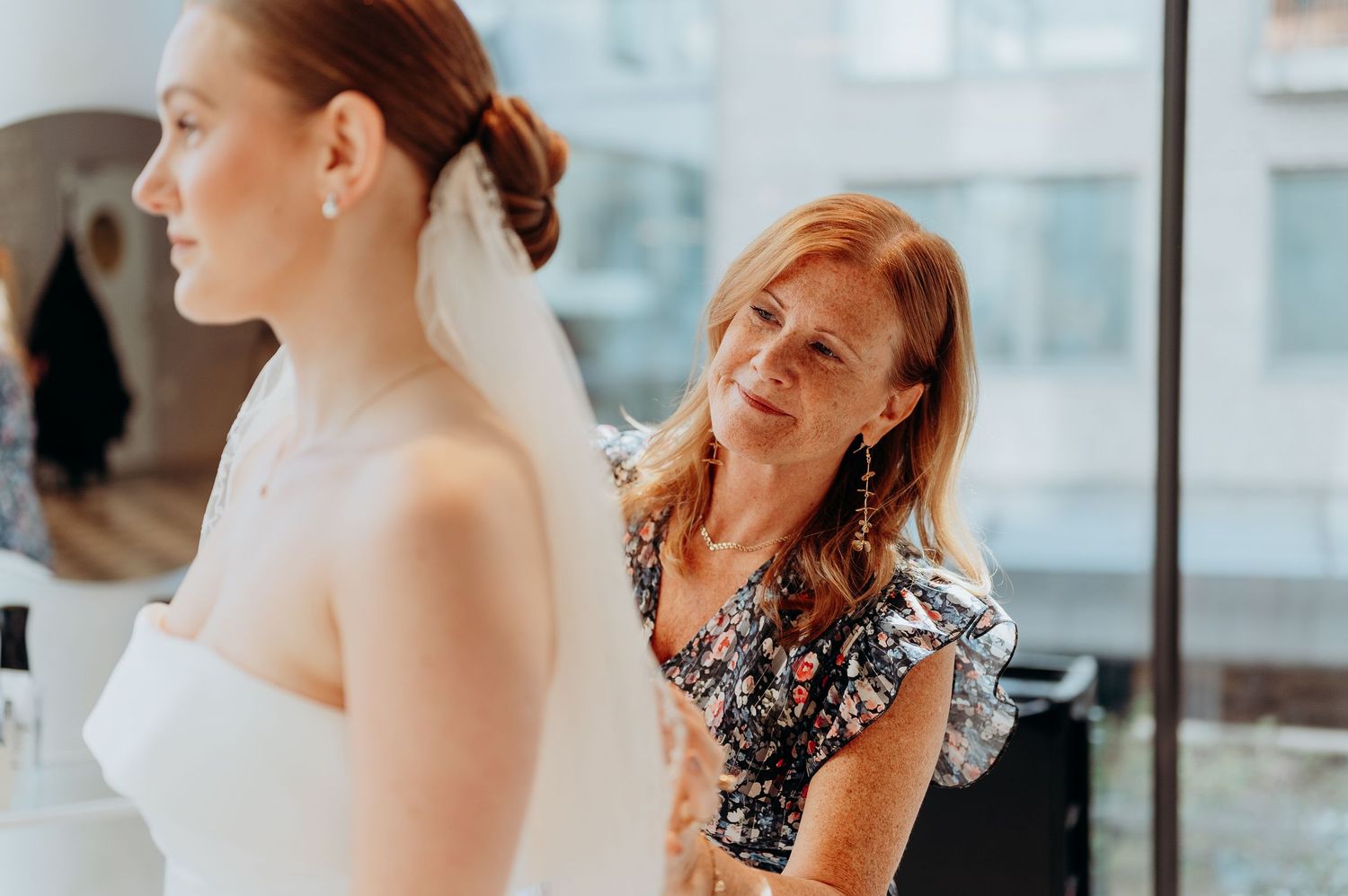 A woman in a floral dress adjusts a bride's white wedding gown and veil near a bright window.