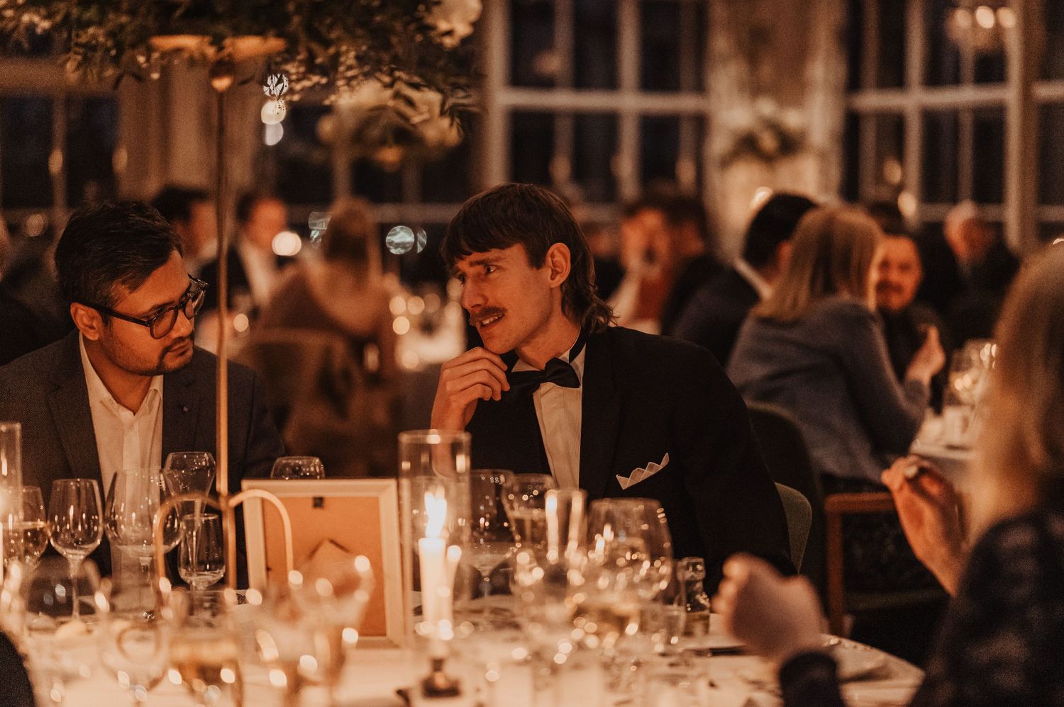 Guests in formal attire enjoying an elegant candlelit dinner reception with glassware and floral arrangements.