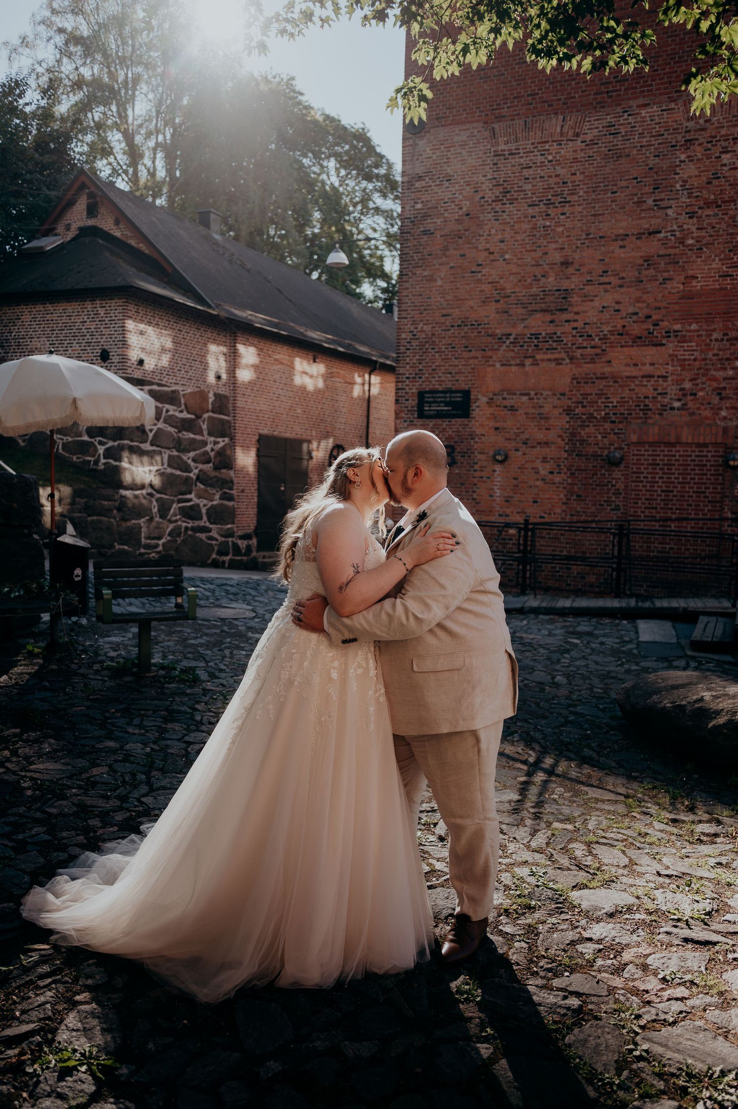 Bride and groom share a romantic kiss in a cobblestone courtyard beside a historic brick building on their wedding day.