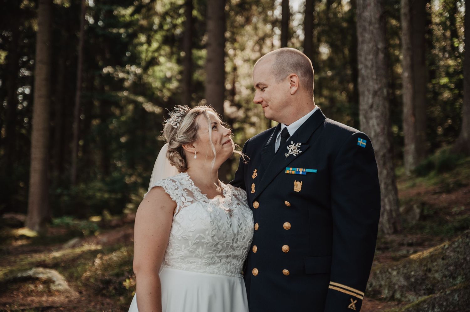Bride and groom share a romantic moment in a forest, with groom in military dress uniform and bride in white lace gown.