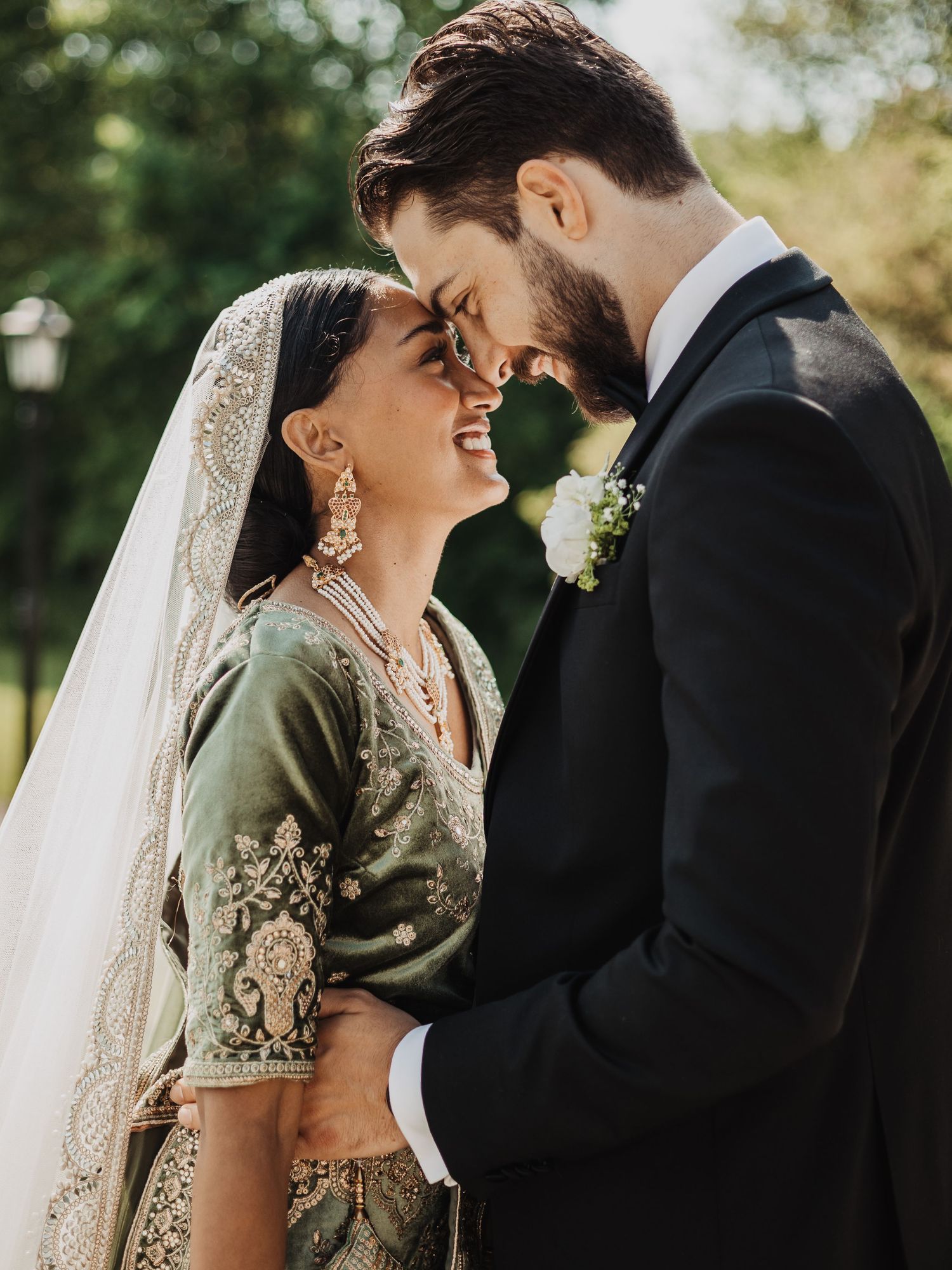 A bride in green embroidered lehenga and groom in black tuxedo share a romantic forehead touch on their wedding day.