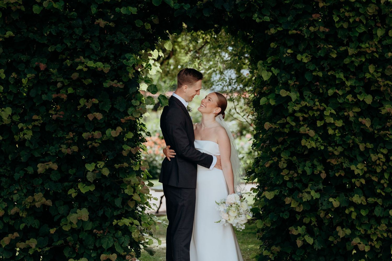 Bride and groom embracing in a stunning ivy-covered archway with a bridal bouquet.