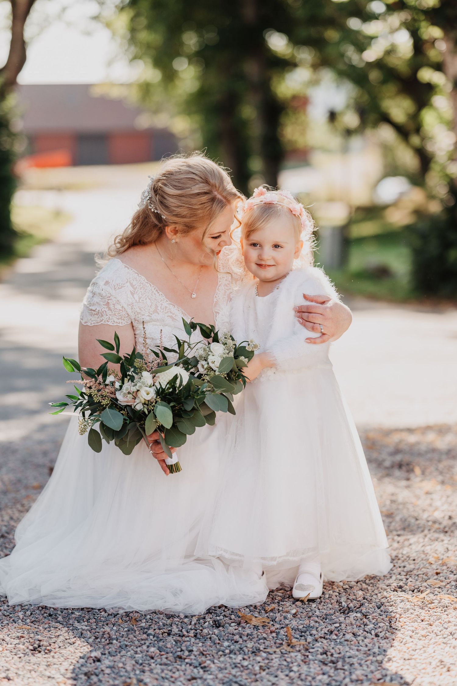 Bride in lace gown kneeling to embrace a smiling flower girl while holding a floral bouquet.