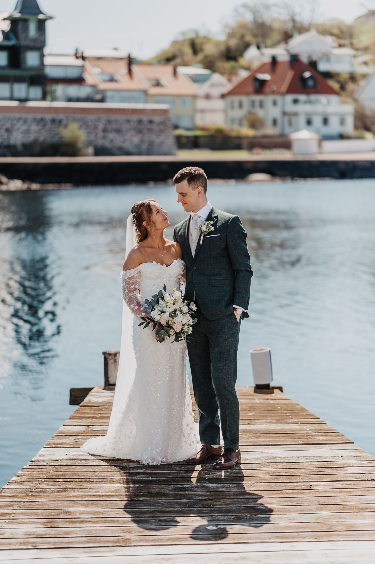 Elegant bride and groom posing on a sunlit waterfront dock with a white floral bouquet.