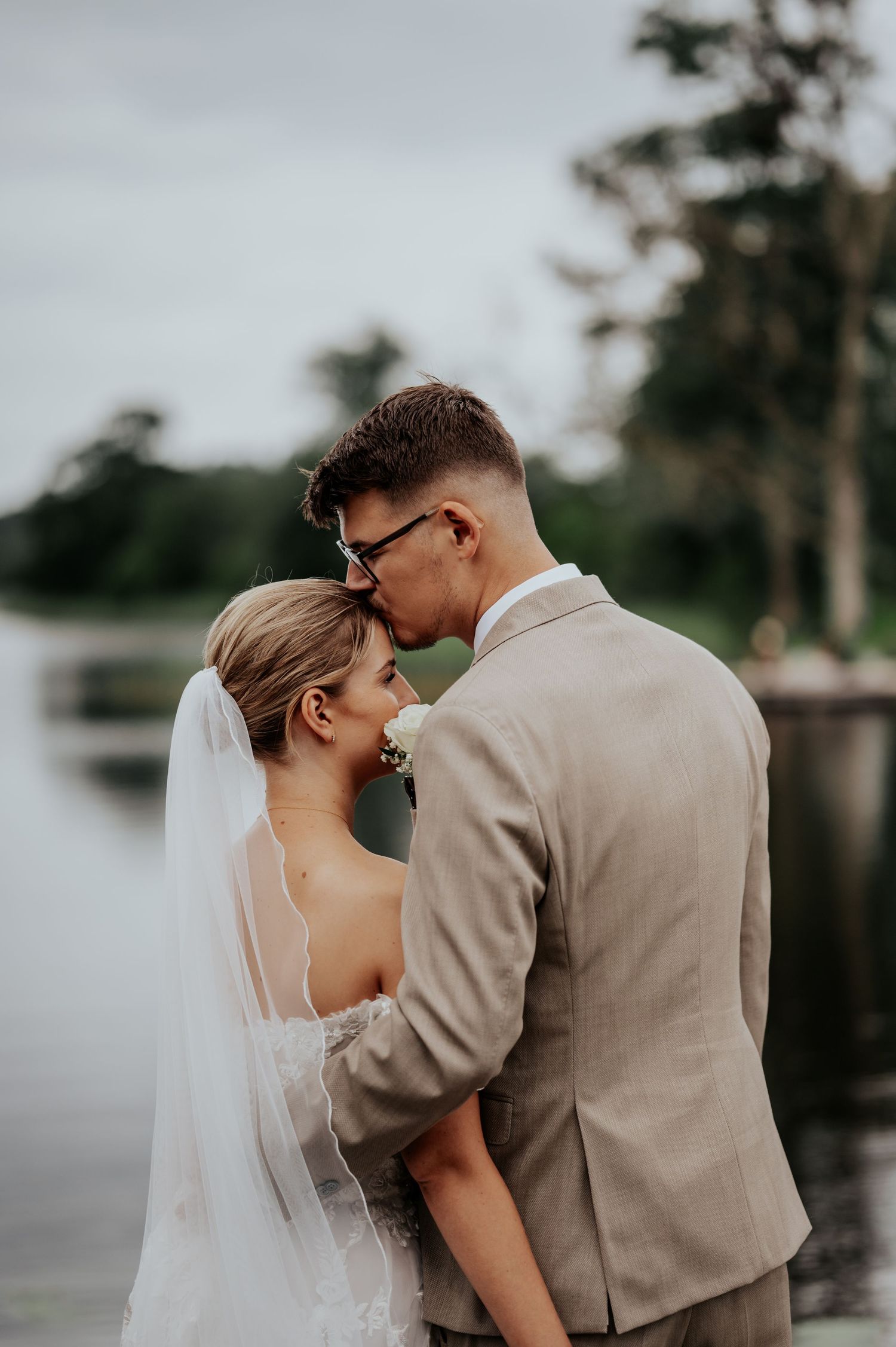 Bride and groom share a tender forehead kiss near a serene lakeside, bride in lace gown with veil, groom in tan suit.