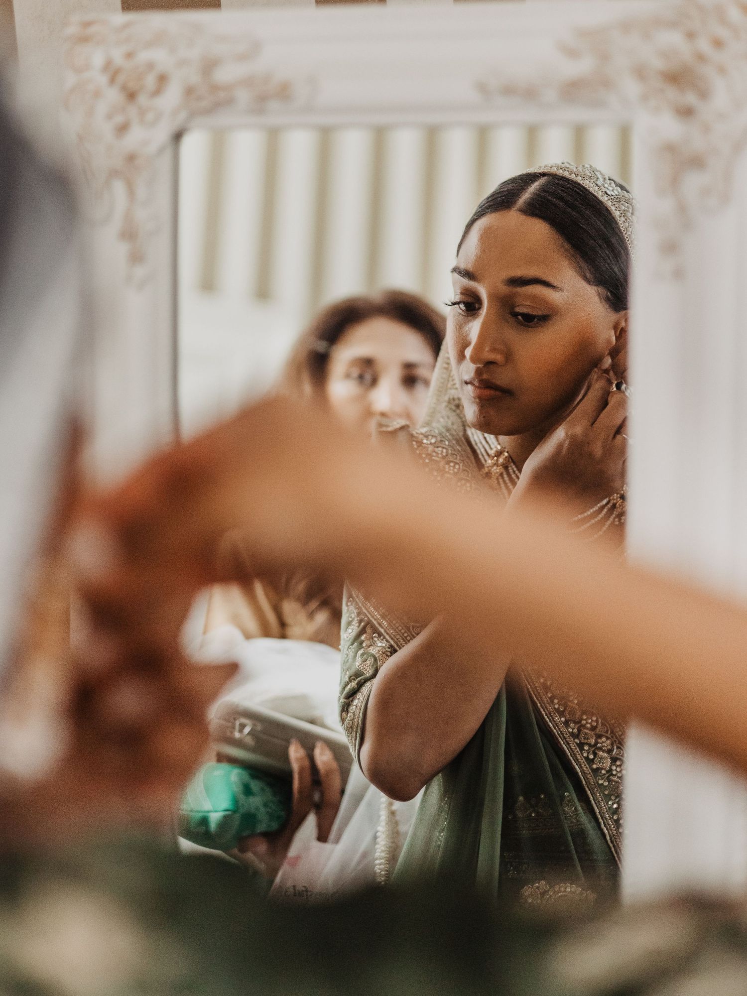 A South Asian bride wearing a tiara adjusts her earring while reflected in an ornate white mirror.