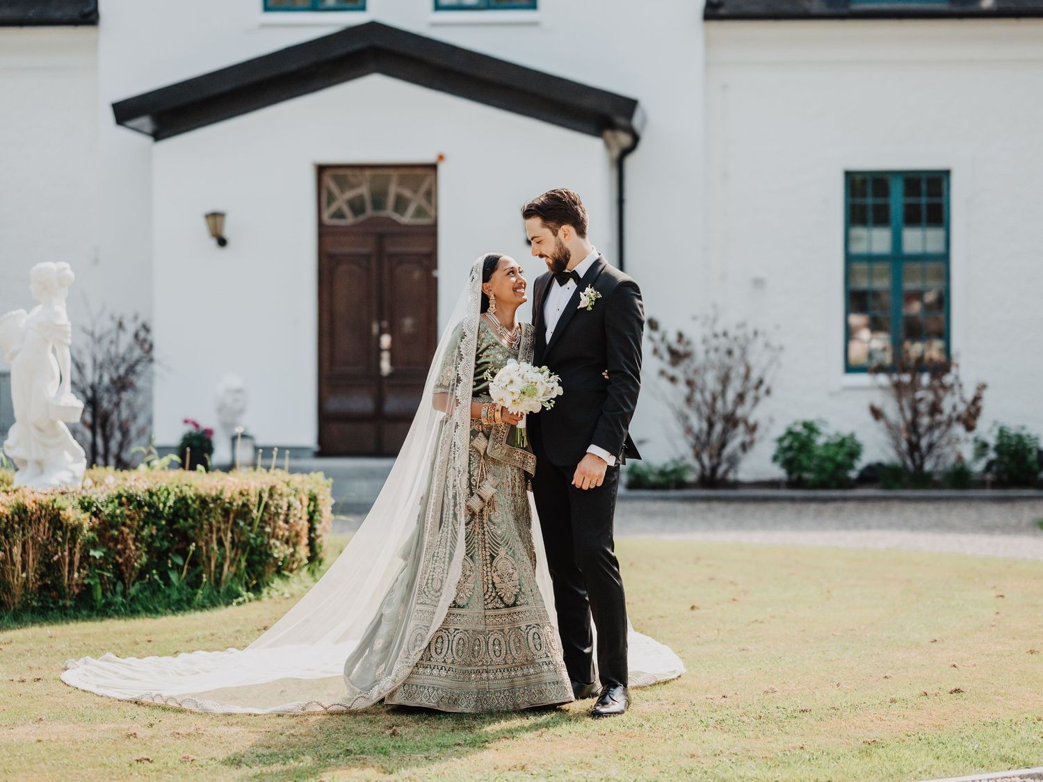 Bride in silver lehenga and groom in black tuxedo share a romantic moment outside a white wedding venue.