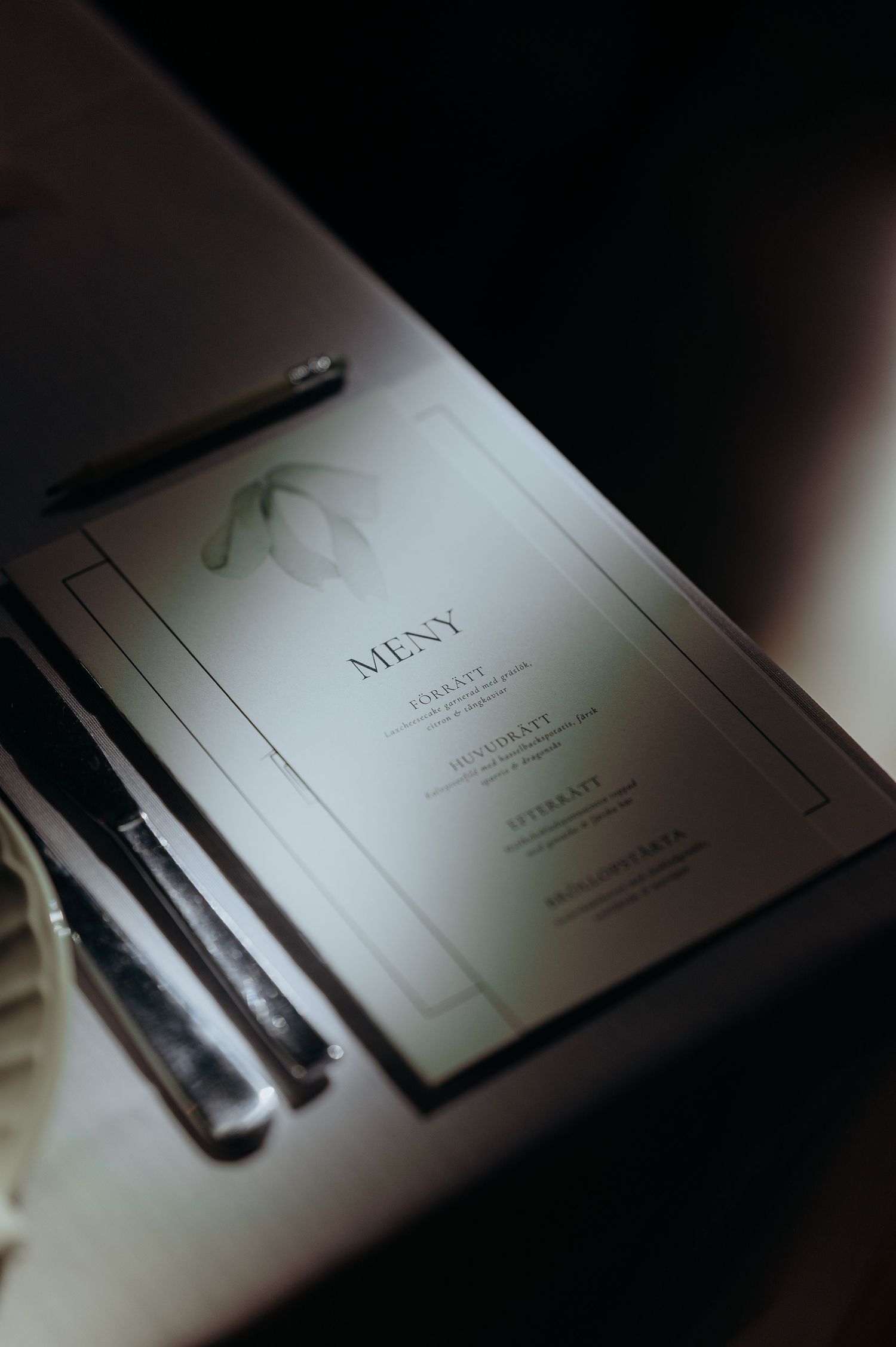 Elegant wedding menu card with botanical snowdrop illustration and silver cutlery on dark table setting.