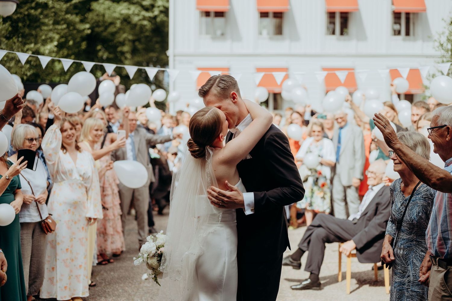 Bride and groom sharing first kiss as newlyweds while guests celebrate with balloons.